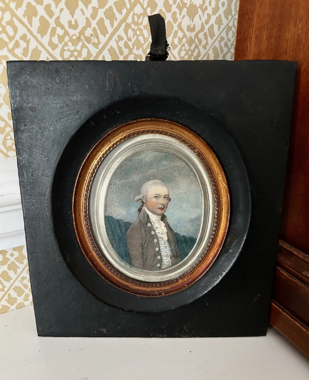 An antique oval watercolour portrait miniature painting of a gentleman wearing a brown coat and wig, housed in a square ebonised frame with a gilt inner slip and hanging loop.