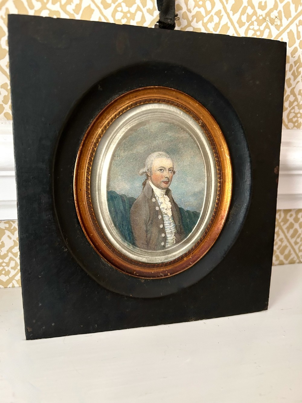 An antique oval watercolour portrait miniature painting of a gentleman wearing a brown coat and wig, housed in a square ebonised frame with a gilt inner slip and hanging loop. View II