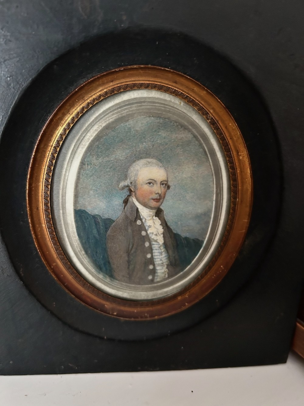 An antique oval watercolour portrait miniature painting of a gentleman wearing a brown coat and wig, housed in a square ebonised frame with a gilt inner slip and hanging loop. View III