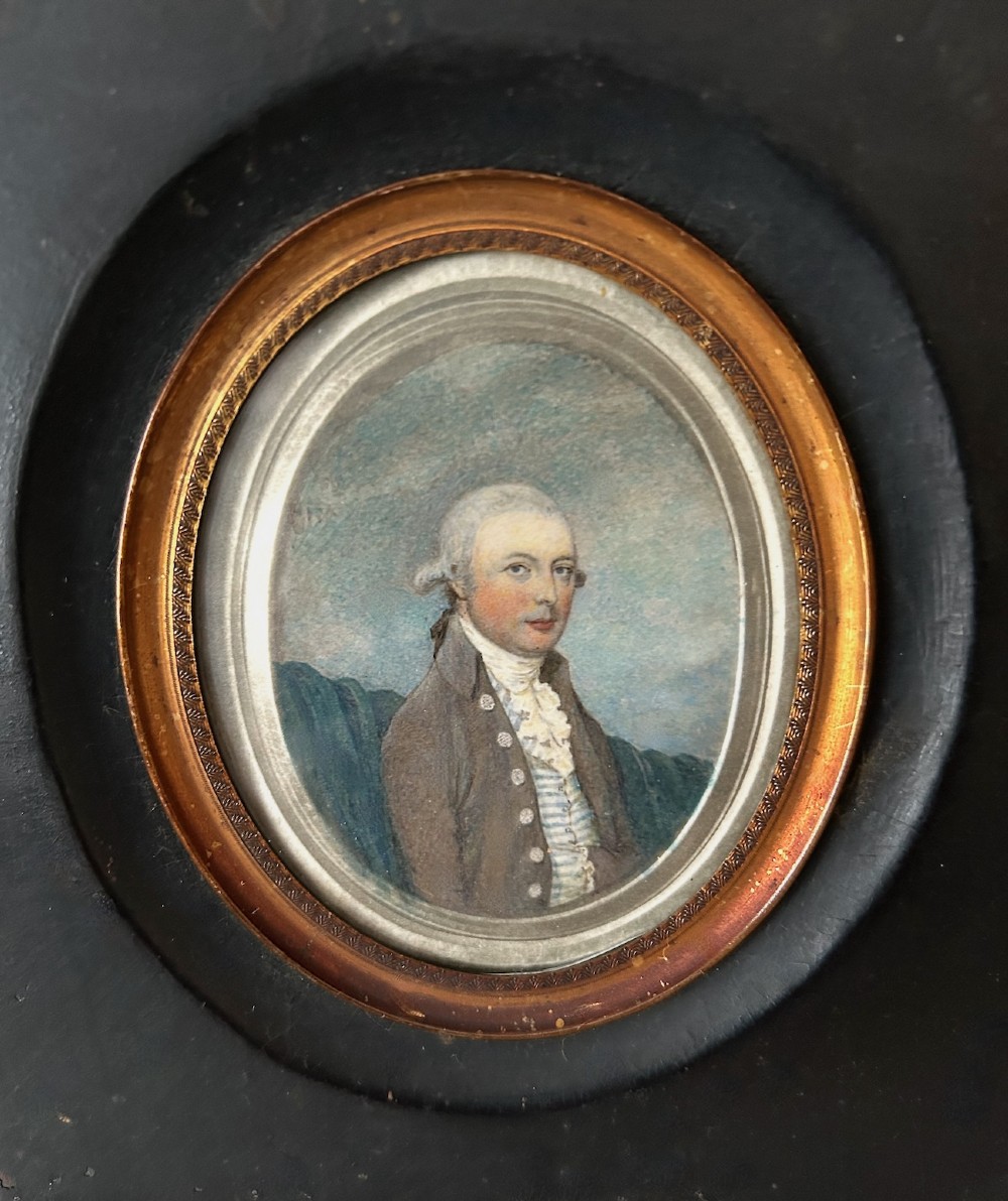 An antique oval watercolour portrait miniature painting of a gentleman wearing a brown coat and wig, housed in a square ebonised frame with a gilt inner slip and hanging loop. View V