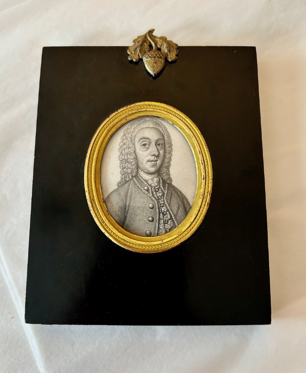 An 18th-century oval monochrome miniature portrait of a gentleman in a period wig, housed in a gilt metal frame mounted on a black ebonised rectangular plaque with an acorn motif hanger.