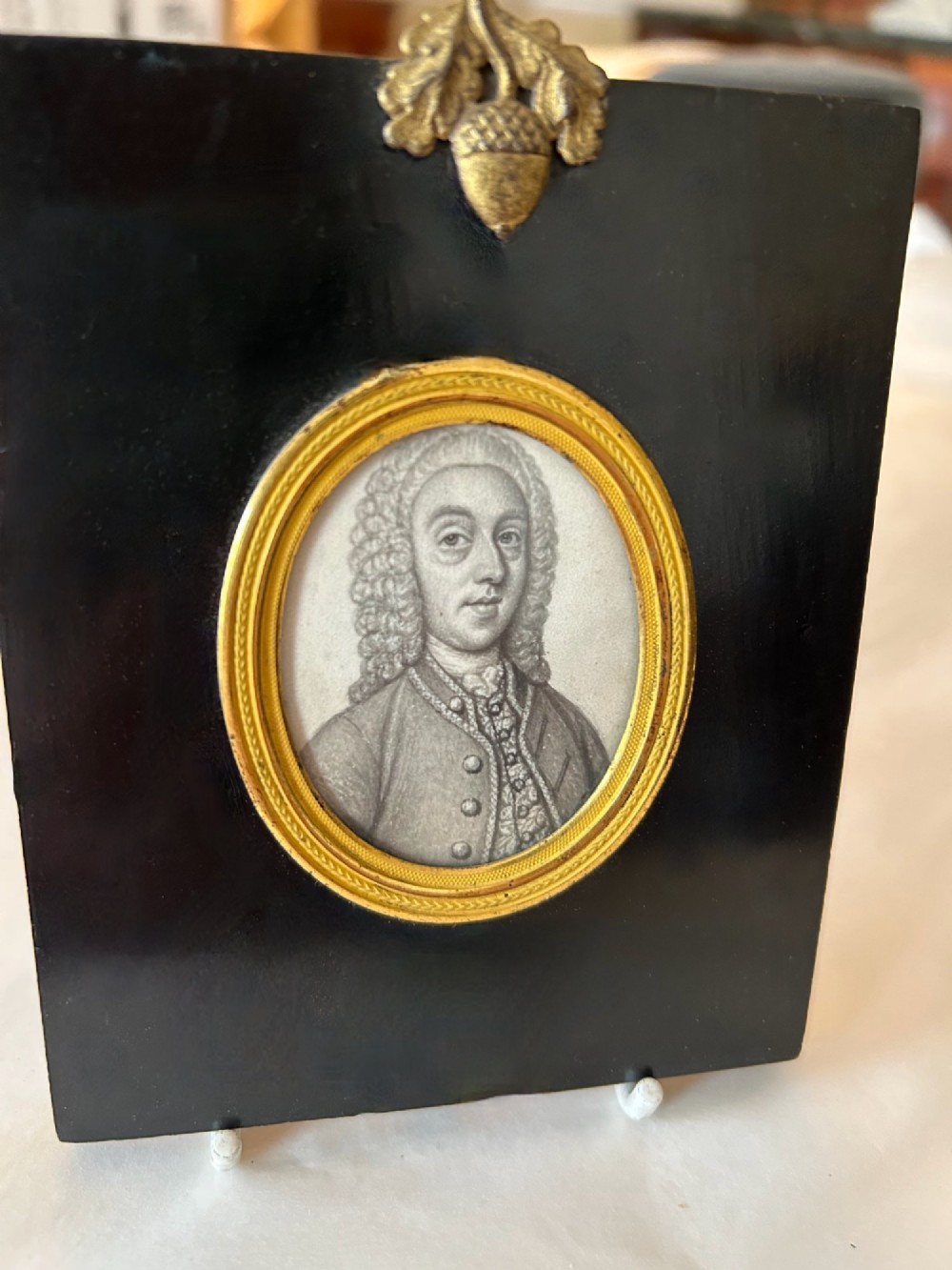 An 18th-century oval monochrome miniature portrait of a gentleman in a period wig, housed in a gilt metal frame mounted on a black ebonised rectangular plaque with an acorn motif hanger. View V