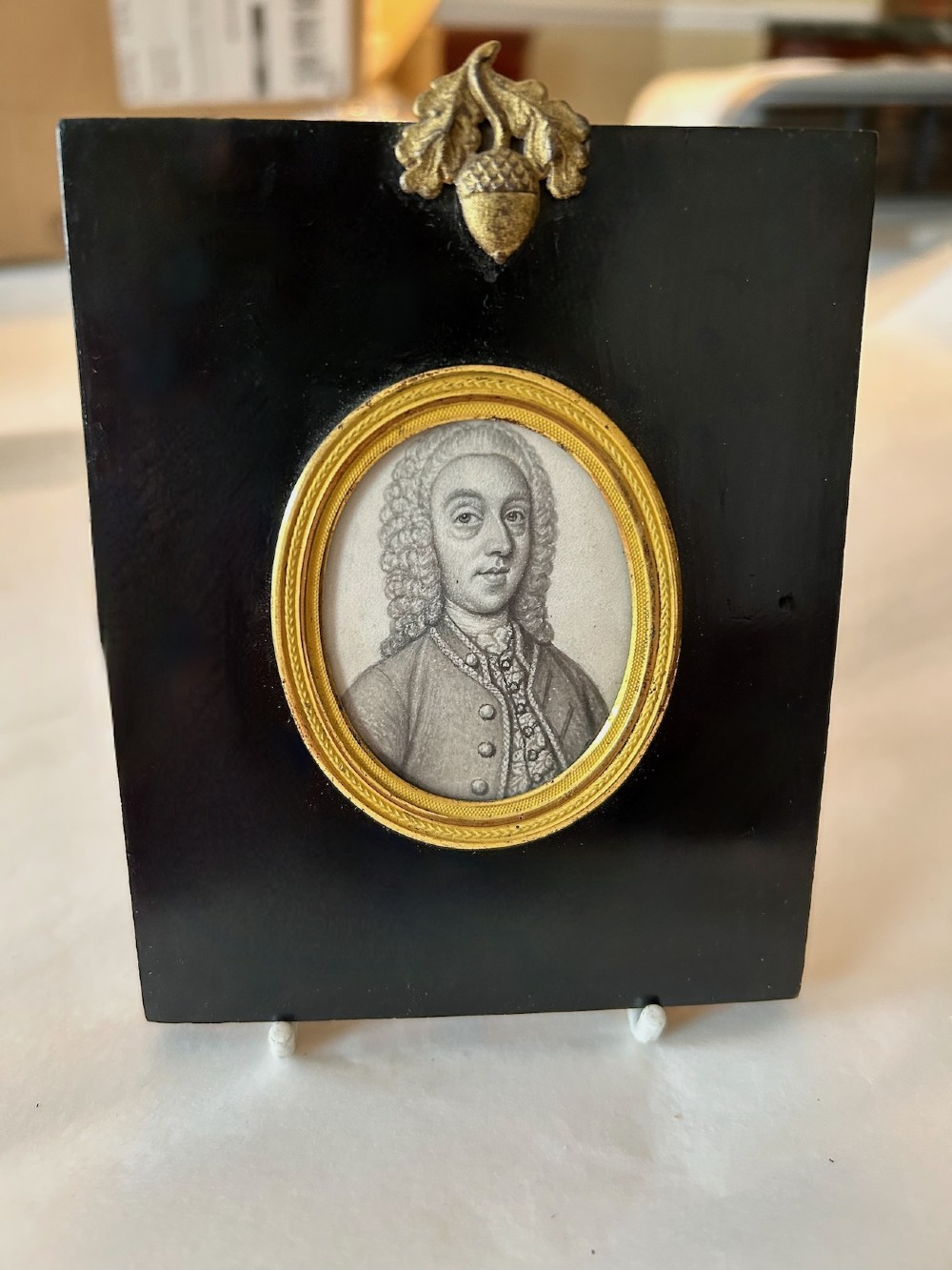 An 18th-century oval monochrome miniature portrait of a gentleman in a period wig, housed in a gilt metal frame mounted on a black ebonised rectangular plaque with an acorn motif hanger. View VI