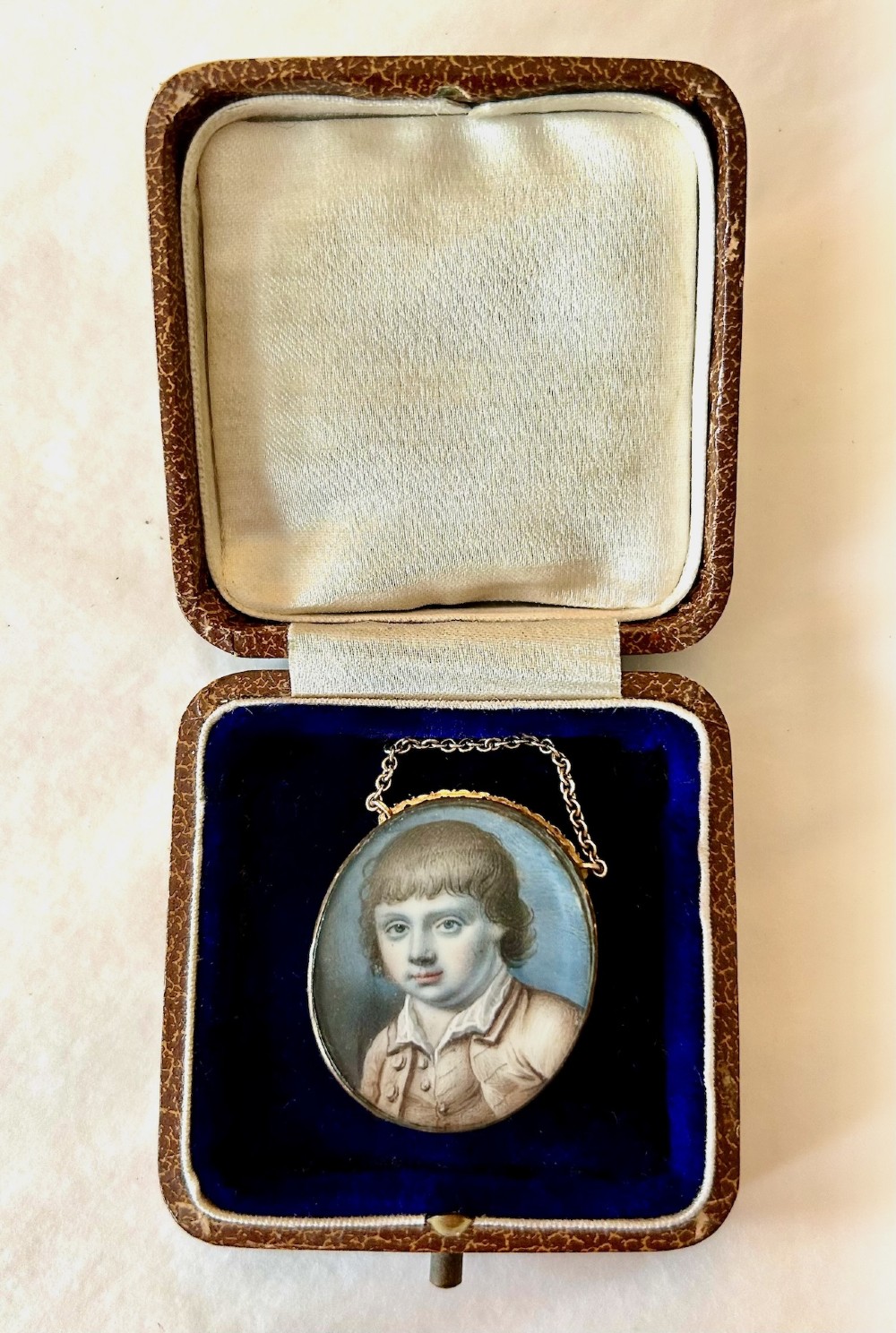 An oval late 18th-century portrait miniature depicting a young boy with light brown hair, set in a gold frame with a chain inside a velvet-lined leather presentation case.