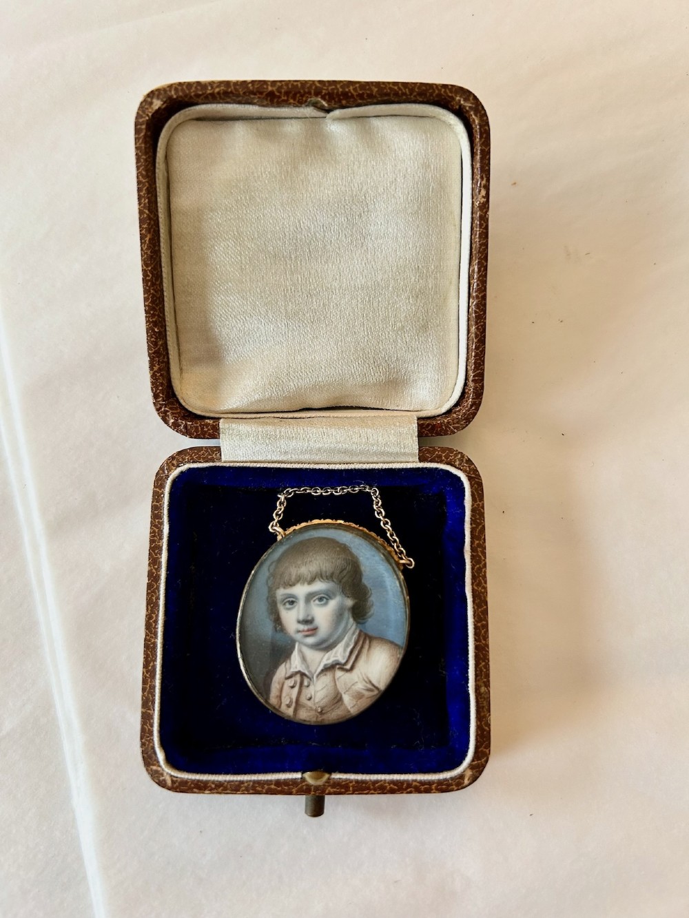 An oval late 18th-century portrait miniature depicting a young boy with light brown hair, set in a gold frame with a chain inside a velvet-lined leather presentation case. View III