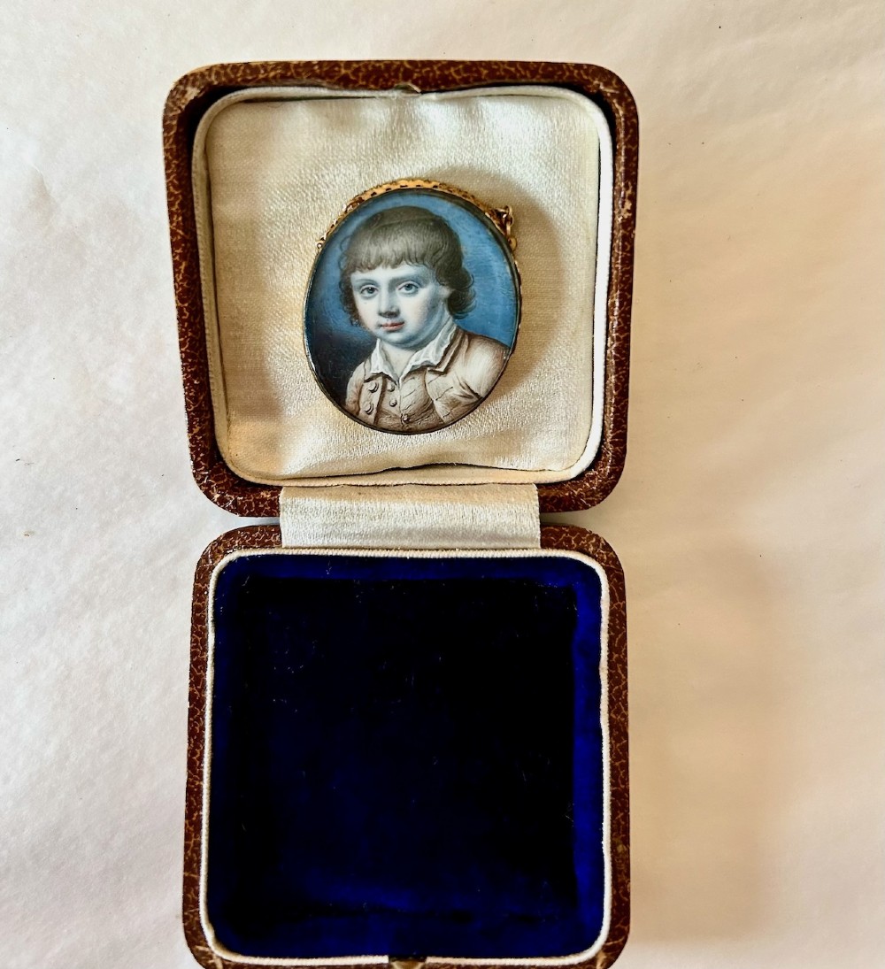 An oval late 18th-century portrait miniature depicting a young boy with light brown hair, set in a gold frame with a chain inside a velvet-lined leather presentation case. View IV