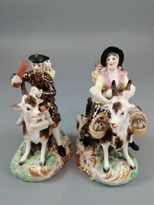 fig 1. A pair of painted porcelain figurines depicting a man and woman in 18th-century attire riding goats with cobalt blue gilded jackets on rococo scroll bases.