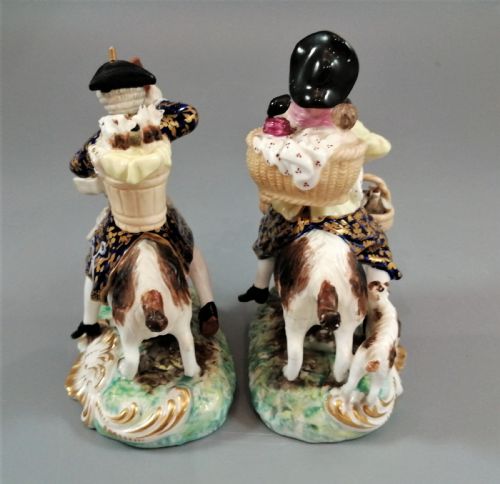 fig 3. A pair of painted porcelain figurines depicting a man and woman in 18th-century attire riding goats with cobalt blue gilded jackets on rococo scroll bases.