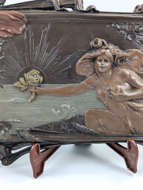 fig 2. A rectangular French Art Nouveau terracotta relief featuring a draped female figure holding a rose against a rising sun background with a metallic finish.