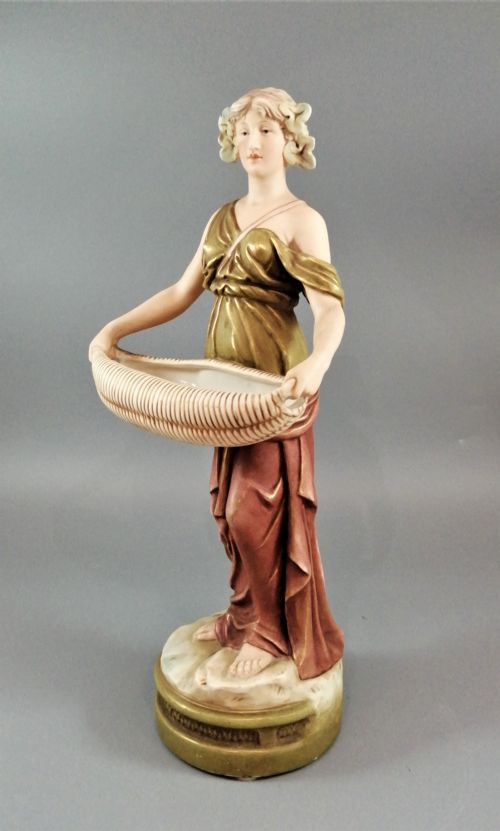 fig 1. An Art Nouveau Royal Dux porcelain figurine depicting a draped maiden standing on a circular gilded pedestal holding a ribbed oval basket.