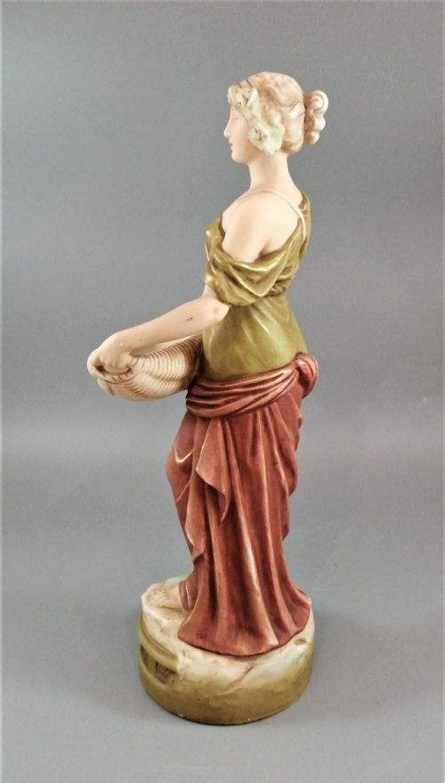 fig 2. An Art Nouveau Royal Dux porcelain figurine depicting a draped maiden standing on a circular gilded pedestal holding a ribbed oval basket.