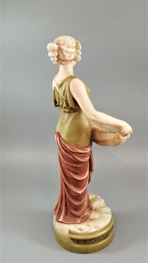 fig 3. An Art Nouveau Royal Dux porcelain figurine depicting a draped maiden standing on a circular gilded pedestal holding a ribbed oval basket.
