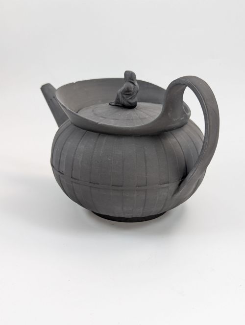 fig 2. An eighteenth-century Wedgwood black basalt teapot featuring a ribbed stoneware body, high loop handle, and a seated female figure finial on the lid.