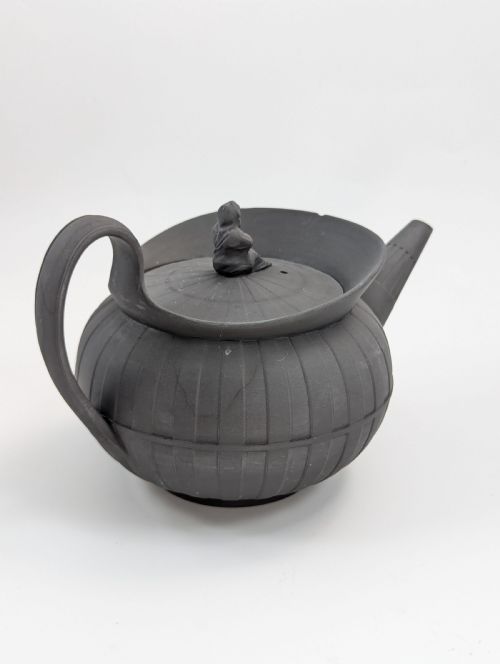 fig 3. An eighteenth-century Wedgwood black basalt teapot featuring a ribbed stoneware body, high loop handle, and a seated female figure finial on the lid.
