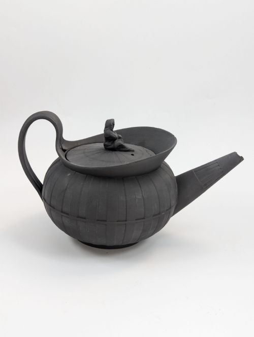 fig 4. An eighteenth-century Wedgwood black basalt teapot featuring a ribbed stoneware body, high loop handle, and a seated female figure finial on the lid.