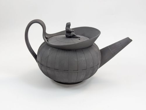 fig 5. An eighteenth-century Wedgwood black basalt teapot featuring a ribbed stoneware body, high loop handle, and a seated female figure finial on the lid.