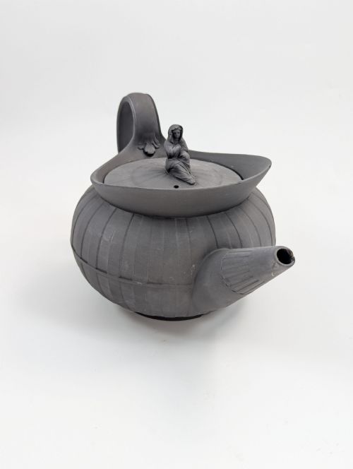 fig 6. An eighteenth-century Wedgwood black basalt teapot featuring a ribbed stoneware body, high loop handle, and a seated female figure finial on the lid.