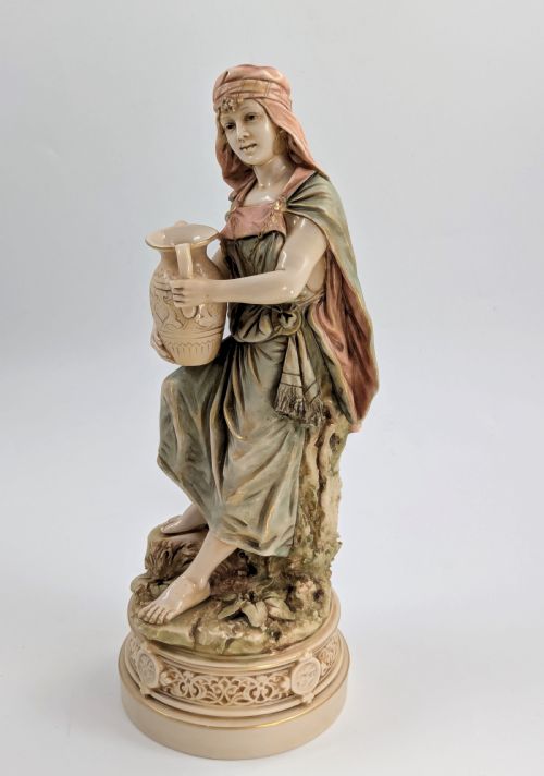 fig 1. A porcelain figurine of a woman in draped robes carrying a double-handled water vessel on a patterned pedestal base.