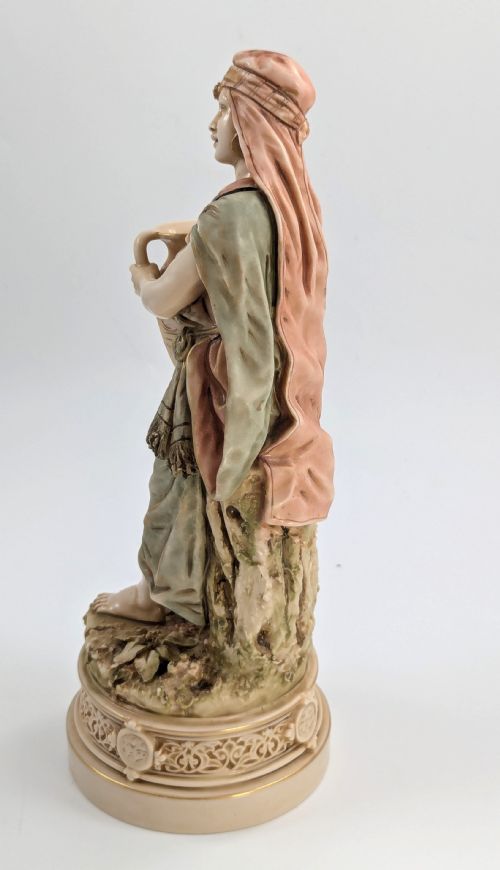 fig 2. A porcelain figurine of a woman in draped robes carrying a double-handled water vessel on a patterned pedestal base.
