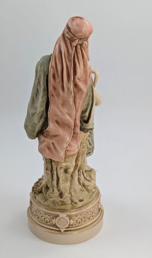 fig 3. A porcelain figurine of a woman in draped robes carrying a double-handled water vessel on a patterned pedestal base.