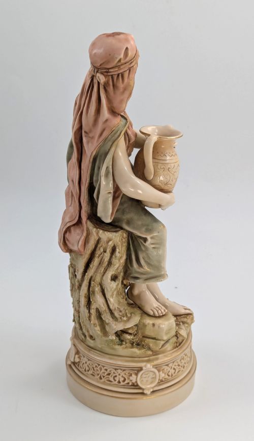 fig 4. A porcelain figurine of a woman in draped robes carrying a double-handled water vessel on a patterned pedestal base.