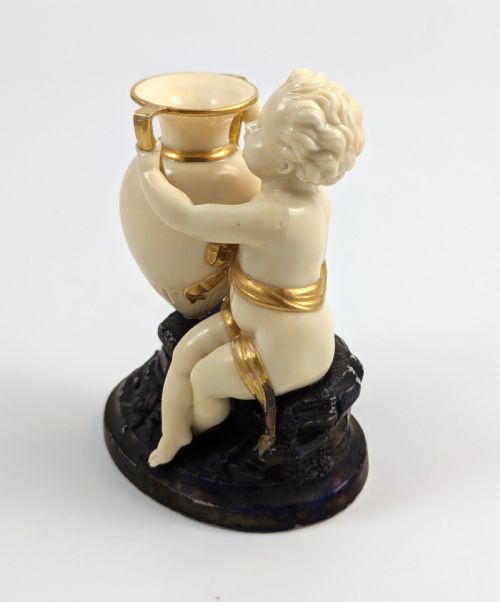 fig 2. An ivory-coloured porcelain figurine depicting a small child seated beside a gilded amphora vessel on a dark base.