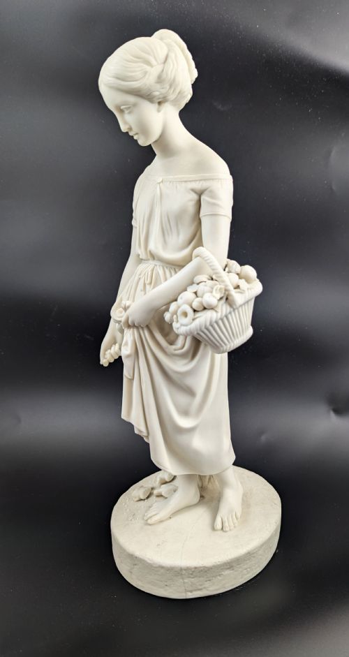 fig 1. An off-white Parian porcelain figurine depicting a standing classical female figure holding a fruit basket on a circular base.
