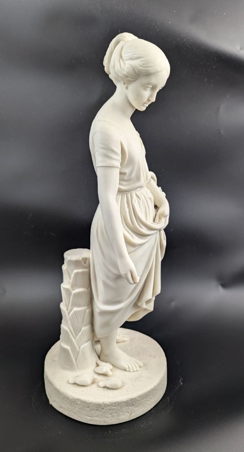fig 5. An off-white Parian porcelain figurine depicting a standing classical female figure holding a fruit basket on a circular base.