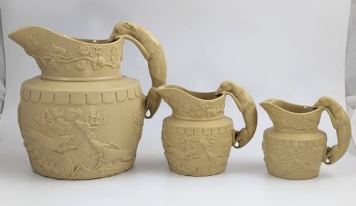 fig 1. A graduated set of three buff yellow Regency ceramic pitchers featuring relief-moulded stag hunting scenes and sculpted hound handles.