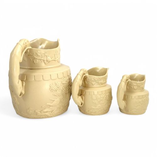 fig 3. A graduated set of three buff yellow Regency ceramic pitchers featuring relief-moulded stag hunting scenes and sculpted hound handles.