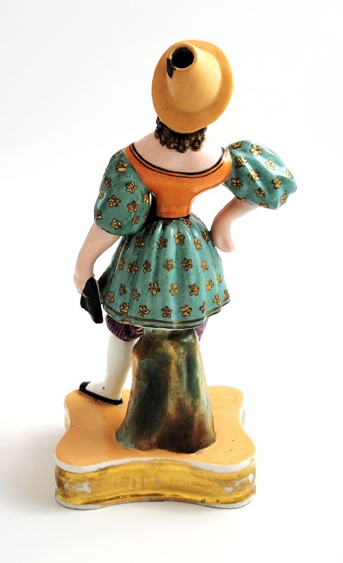 fig 1. An early 19th-century French hard-paste porcelain figural scent bottle depicting a standing woman in a teal patterned dress, orange bodice, and yellow hat, mounted on a gilded shaped base.
