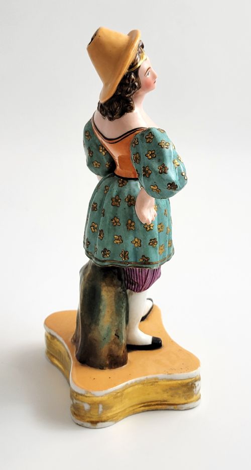 fig 2. An early 19th-century French hard-paste porcelain figural scent bottle depicting a standing woman in a teal patterned dress, orange bodice, and yellow hat, mounted on a gilded shaped base.
