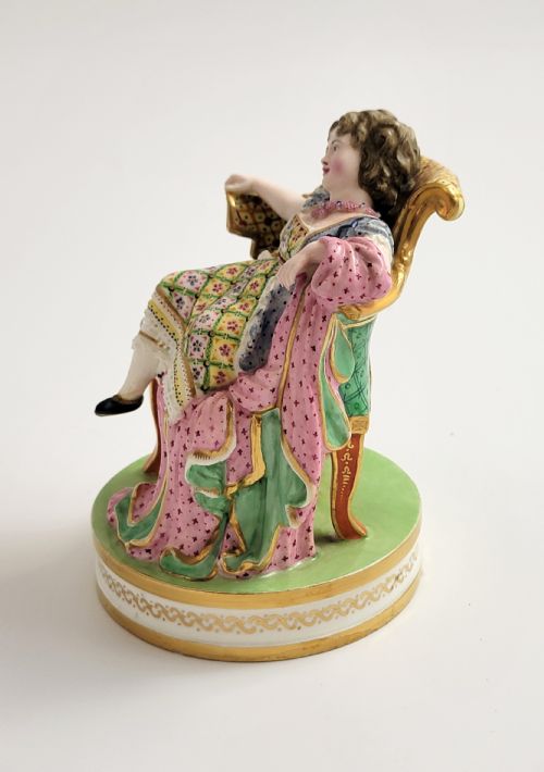 fig 1. An 18th-century Derby soft-paste porcelain figurine depicting a seated woman in a colourful dress reclining on a gilded armchair.