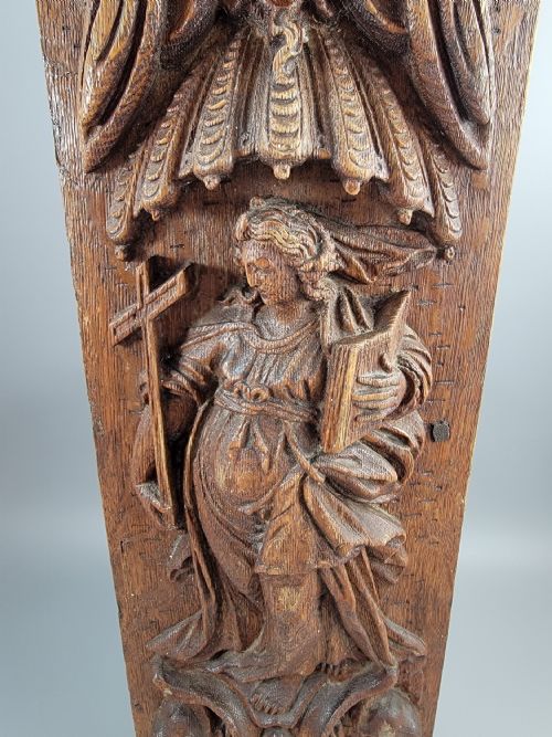 fig 1. A vertical carved oak panel displaying a high-relief figure of Faith holding a cross and a book beneath ornate Baroque scrollwork.
