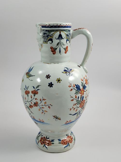 fig 1. An eighteenth-century tin-glazed earthenware jug featuring a moulded mask spout, loop handle, and hand-painted polychrome floral and bird motifs on a white background.
