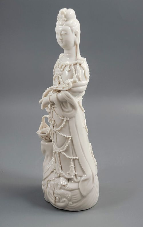 fig 1. An early 20th-century Blanc de Chine porcelain figure of Guanyin wearing beaded robes and holding a fish basket on a wave pedestal.