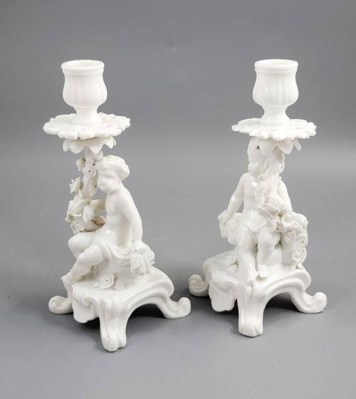 fig 1. A pair of white glazed porcelain candlesticks featuring sculpted putti figures resting against floral encrusted columns on scrollwork bases.