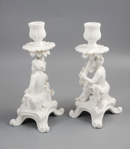 fig 2. A pair of white glazed porcelain candlesticks featuring sculpted putti figures resting against floral encrusted columns on scrollwork bases.