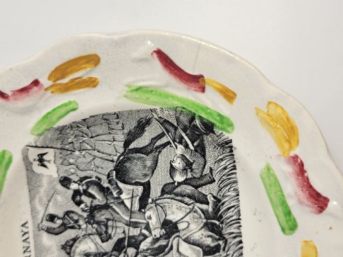 fig 1. A circular Victorian ceramic plate featuring a central black transfer print of the Battle of Tchernaya surrounded by a scalloped rim with green, red, and yellow enamel accents.