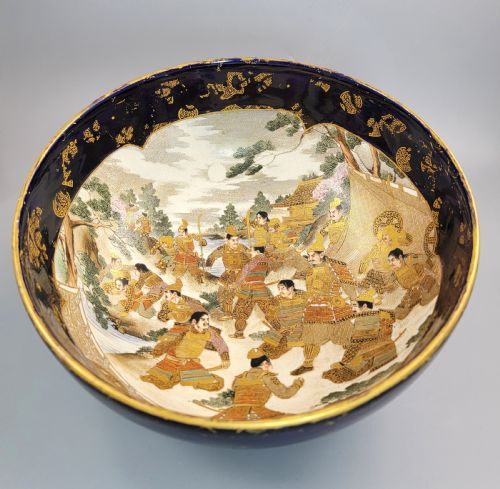 fig 1. A circular Japanese Satsuma earthenware bowl featuring a central scene of samurai warriors in gold and polychrome enamels surrounded by a dark cobalt blue rim decorated with intricate gold scrollwork.