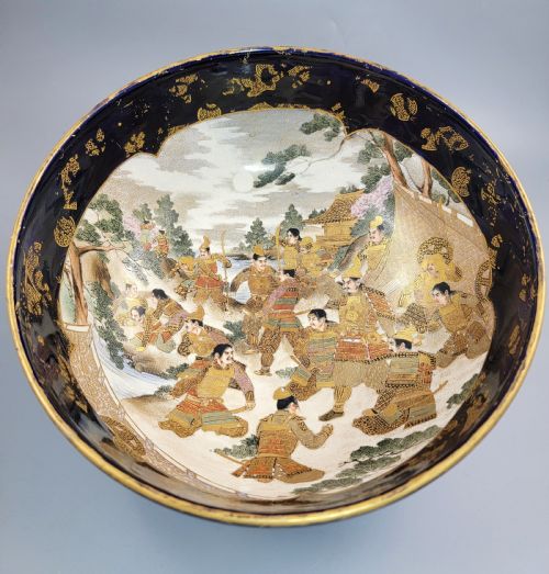 fig 2. A circular Japanese Satsuma earthenware bowl featuring a central scene of samurai warriors in gold and polychrome enamels surrounded by a dark cobalt blue rim decorated with intricate gold scrollwork.
