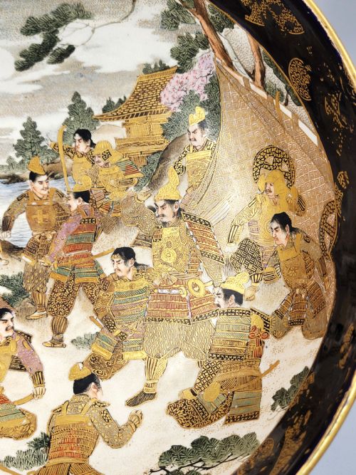 fig 4. A circular Japanese Satsuma earthenware bowl featuring a central scene of samurai warriors in gold and polychrome enamels surrounded by a dark cobalt blue rim decorated with intricate gold scrollwork.