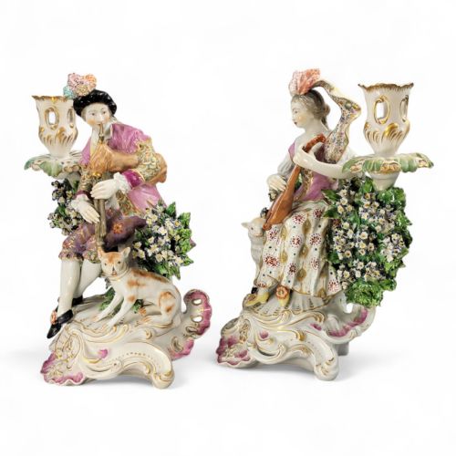 fig 1. A pair of 18th-century Derby porcelain candlestick figures depicting a male bagpipe player with a dog and a female mandolin player with a lamb, set against floral boscage backgrounds on gilded scrollwork bases.