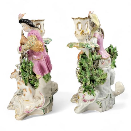 fig 2. A pair of 18th-century Derby porcelain candlestick figures depicting a male bagpipe player with a dog and a female mandolin player with a lamb, set against floral boscage backgrounds on gilded scrollwork bases.