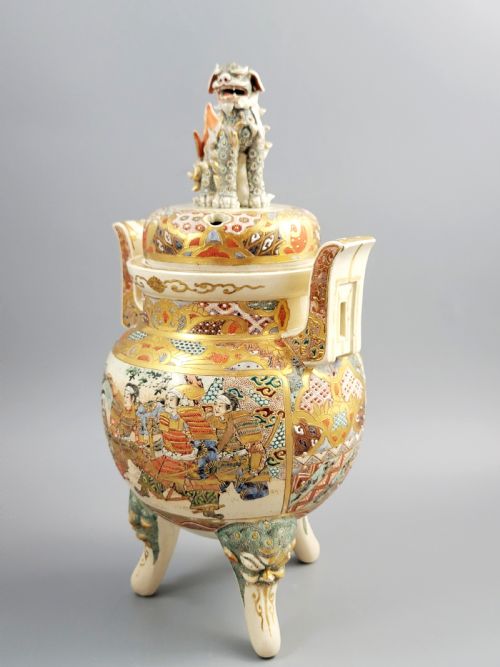 fig 1. A cream-coloured Japanese Satsuma earthenware incense burner featuring a lid with a shishi lion finial, upright handles, tripod feet, and detailed polychrome paintings of samurai warriors with gold accents.