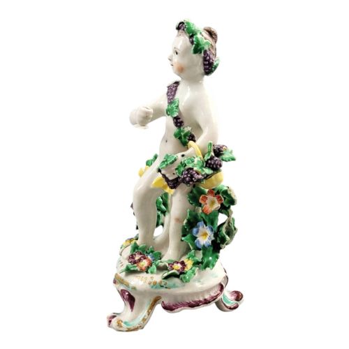 fig 1. An 18th-century Bow soft-paste porcelain figure of a putto adorned with grapevines, standing on a Rococo scroll base with colourful floral bocage behind.