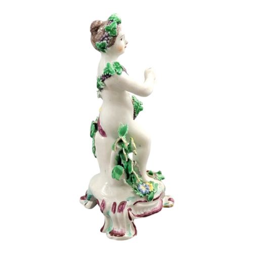 fig 3. An 18th-century Bow soft-paste porcelain figure of a putto adorned with grapevines, standing on a Rococo scroll base with colourful floral bocage behind.