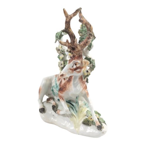 fig 1. An 18th-century soft-paste porcelain figurine depicting a recumbent brown and white spotted stag resting in front of a leafy bocage with small yellow flowers.