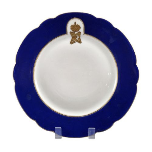 fig 1. A white round porcelain plate featuring a thick cobalt blue scalloped border and a gilded Imperial monogram topped with a crown.