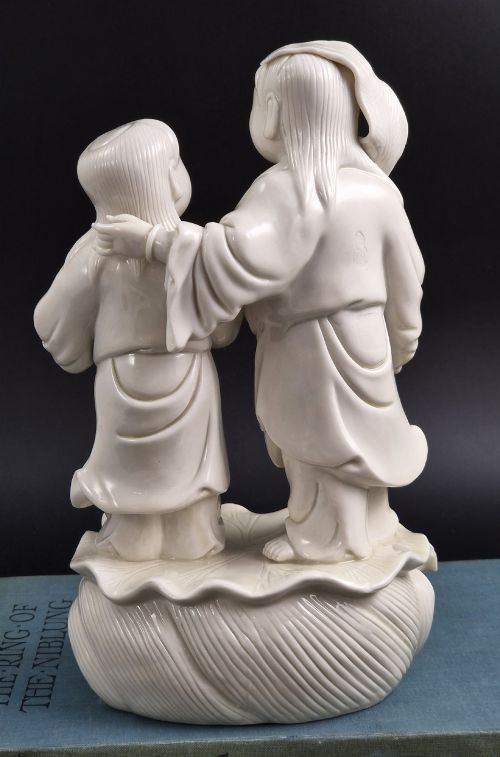 fig 5. A cream white glazed porcelain figure group featuring two standing figures positioned on a lotus leaf and wave carved base.